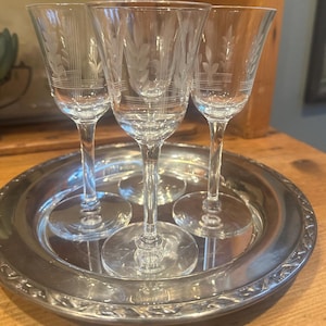 May include: Three clear, stemmed wine glasses with etched leaf designs are arranged on a silver-colored serving tray. The glasses have a classic shape with a slender stem and a flared bowl. The tray has a decorative edge.