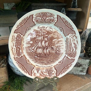 Könnte beinhalten: Eine braun-weiße Vintage-Platte mit einer detaillierten Illustration eines Segelschiffs und dem Text "Friendship of Salem". Die Platte wird auf einem dekorativen Messingständer präsentiert, der ihr aufwendiges Design und ihr historisches Thema hervorhebt. Durchmesser: ca. 25 cm.