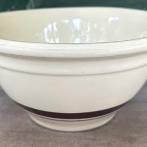 May include: A cream-colored ceramic mixing bowl with a smooth, glossy finish and a thick, dark brown horizontal band encircling the lower portion of the exterior. The bowl features a wide, rounded rim and a deep, open interior basin.