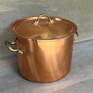 May include: A polished copper stockpot with a matching lid and two brass side handles. The pot features a cylindrical body and a rounded lid with a central brass handle. The metallic surface reflects light, showing a warm, reddish-brown hue.
