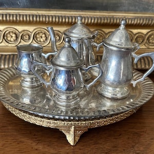 May include: A miniature silver-toned tea set consisting of a lidded teapot, a lidded sugar bowl, and a small creamer, all arranged on a round, scalloped-edge silver-toned tray with a decorative gold-toned base featuring three ornate feet.