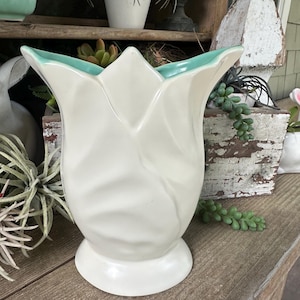 Könnte beinhalten: Eine weiße, tulpenförmige Keramikvase mit türkisfarbenem Rand. Die Vase hat eine breite, ausgestellte Öffnung und einen runden Sockel. Die Vase steht auf einer Holzoberfläche. Dekorative Wohnkultur.