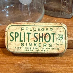 May include: A rectangular metal tin for Pflueger Split Shot Sinkers with a cream-coloured background and green text. The label features the text PFLUEGER SPLIT SHOT SINKERS, TRADE MARKS REG. U.S. PAT. OFFICE, MADE IN U.S.A., and a small dog illustration.