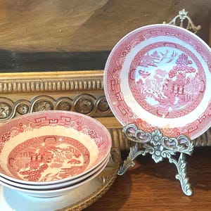 May include: A set of ceramic bowls featuring a red and white transferware pattern depicting a traditional willow scene with pagodas, trees, and birds. One bowl is displayed upright on a metal stand, while three others are stacked below it.