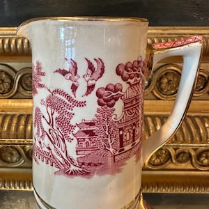 May include: A ceramic pitcher featuring a cream-colored base with a deep red transferware pattern depicting a landscape with pagodas, willow trees, and flying birds. The rim and handle are accented with gold trim, and the handle has a red pattern.