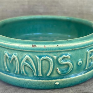 Puede incluir: Un cuenco de cerámica turquesa con una forma ancha y redondeada. El exterior presenta letras en relieve que dicen "D. MANS. BEG." El cuenco tiene un aspecto envejecido y desgastado.