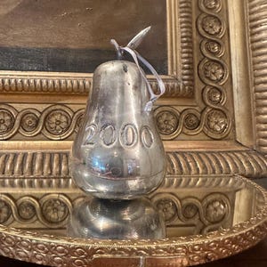 May include: A silver-toned pear-shaped ornament featuring the embossed number 2000 on its front surface. The object has a smooth, reflective metallic finish and includes a thin, light-colored ribbon loop attached to the top stem for hanging purposes.