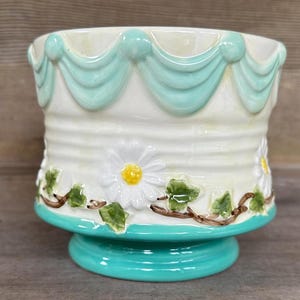 May include: A ceramic planter featuring a cream-colored body with a raised light blue scalloped swag design around the top rim. The exterior is decorated with embossed white daisy flowers and green ivy vines, finished with a solid teal base.
