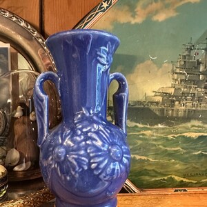 May include: A glossy cobalt blue ceramic vase featuring two side handles and a raised floral relief pattern on the front. The vase has a flared rim, a rounded body, and a circular base, showcasing a vibrant monochromatic finish throughout the piece.