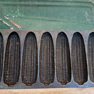 May include: A dark gray cast iron cornbread pan featuring seven individual molds shaped like ears of corn. The rectangular pan has integrated handles on both ends and a textured interior surface designed to create the detailed pattern of corn kernels.