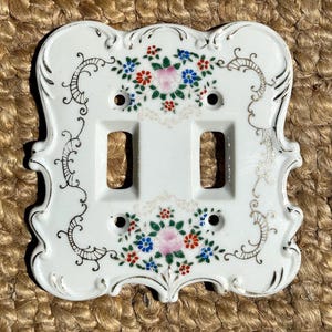 Peut inclure: Une plaque d'interrupteur double en céramique blanche avec un bord festonné et une bordure dorée. La surface présente des motifs floraux peints à la main en rose, bleu, vert et rouge, disposés au-dessus et au-dessous des deux ouvertures.
