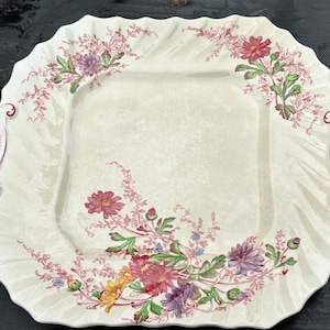 May include: A square, off-white serving platter with a scalloped edge and two handles. The platter is decorated with floral patterns in shades of pink, purple, yellow, and green. The design is symmetrical, with floral arrangements in each corner.