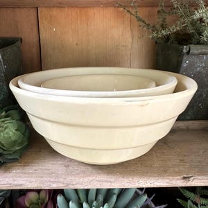 May include: A set of two nested cream-coloured ceramic mixing bowls with a smooth, glossy finish. Each bowl features a wide, flared rim and two distinct horizontal ridges encircling the exterior, creating a tiered, rounded profile.