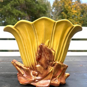 May include: A yellow ceramic vase with a fluted design. The vase has a brown and gold floral accent on the front. The vase is sitting on a dark surface.