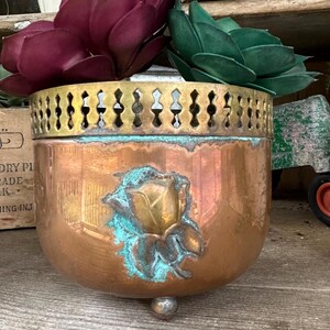May include: A copper-colored planter with a decorative brass rim and a rose emblem. The planter holds two faux succulents, one deep burgundy and the other green. The planter has a small, round foot.