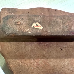 May include: A worn wooden block with a handle, showing signs of age and use. The block is a reddish-brown color, with a textured surface and visible wood grain. A small sticker is affixed to the handle.
