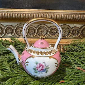 Op de afbeelding: Een kleine porseleinen theepot met een wit en roze kleurenschema en gouden randen. De behuizing is versierd met een handgeschilderde roze roos en blauwe bloemaccenten. Inclusief een bijpassend roze deksel met gouden knop en gouden handvat.