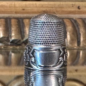Könnte beinhalten: Ein silberfarbener Fingerhut aus Metall mit einer texturierten, genoppten Kuppeloberseite zum Schutz der Nadel und einem glatten, polierten Band an der Basis. Der untere Teil ist mit geprägten, erhabenen Schnörkelmustern verziert.