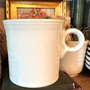 Może przedstawiać: Biały ceramiczny kubek z zaokrąglonym uchwytem. Kubek ma cylindryczny kształt i lekko rozszerzany brzeg. Kubek stoi na ciemnozielonej książce. Z tyłu znajduje się obraz w złotej ramie.