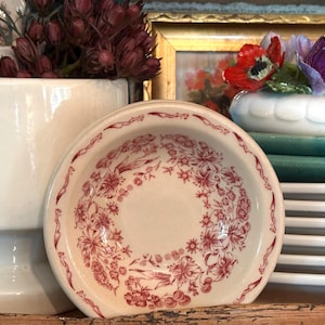 May include: A small, round, cream-colored ceramic saucer featuring a detailed red floral transferware pattern. The design includes a central ring of various blossoms and leaves, with a decorative border pattern along the outer rim of the dish.