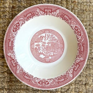 May include: A round ceramic bowl featuring a red and white transferware pattern. The center displays a detailed landscape with a pagoda, bridge, and birds, while the wide rim is decorated with an intricate geometric and floral border design.