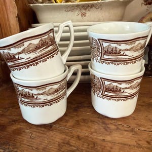 May include: Four white ceramic cups featuring a brown transferware landscape pattern depicting mountains, trees, and a lake. Each cup has a distinct angular handle and a decorative border surrounding the central scene, arranged in two stacks.