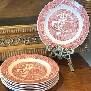 May include: A set of five round ceramic plates featuring a red and white transferware pattern. The design depicts a detailed landscape with birds, trees, and architectural structures. One plate is displayed upright on a metal stand, while four plates are stacked horizontally below it.