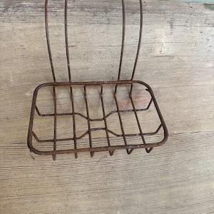 May include: A rusty, vintage metal soap dish with a rectangular basket and curved hanging hooks. The soap dish is made of thin, welded metal rods, showing signs of age and wear. The design is simple and functional.