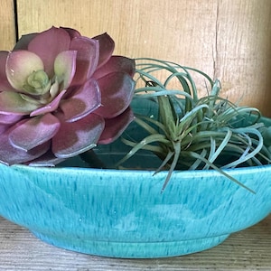 May include: A shallow, turquoise ceramic bowl with a speckled glaze finish. The bowl contains a large, dark purple artificial succulent, a spiky green air plant, and a trailing string of small, light green artificial beads hanging over the side.