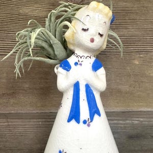 May include: A vintage ceramic figurine of a girl in a white dress with blue accents, holding a plant. The figurine has a crackle finish and is decorated with small purple flowers. The girl's hair is blonde, and she has closed eyes.