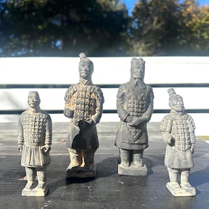 May include: Four small, weathered statues of ancient Chinese terracotta warriors. Each figure is depicted in a standing pose, wearing detailed armor and traditional headwear. The statues are various sizes and have a muted, earthy color palette.