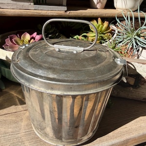 May include: A small, round, galvanized metal bucket with a matching lid and a sturdy, arched wire handle. The container features a vertical ribbed texture along its sides and a secure latch mechanism on the side of the lid for closure.