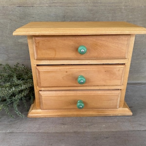 May include: A small, wooden jewelry box with three drawers. The box is light brown with green knobs. The jewelry box is sitting on a wooden surface.
