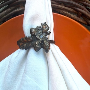 Puede incluir: Un servilletero metálico con un diseño detallado en tono bronce de hojas de roble y bellotas. El anillo está envuelto alrededor de una servilleta de tela blanca lisa, mostrando la textura intrincada y los elementos botánicos esculpidos.