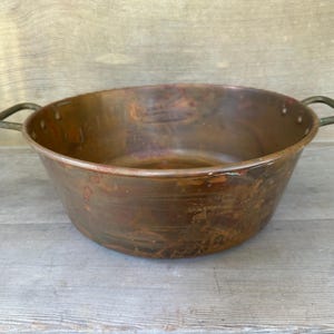 May include: A round, wide-mouthed copper basin featuring a polished metallic finish with visible signs of natural oxidation and patina. The vessel includes two sturdy, riveted metal handles attached to opposite sides of the upper rim.