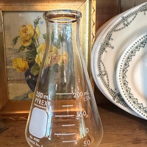 May include: A clear Pyrex Erlenmeyer flask, capacity 250 ml, with graduated markings. The flask has the text "PYREX Made in Germany" and "No. 4980 STOPPER No. 6". The flask is displayed near a framed painting and decorative plates.