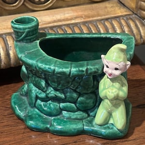 May include: A vintage ceramic planter in the shape of a well, painted in shades of green. A small, smiling elf figure is kneeling beside the well. The planter has a textured, stone-like appearance.