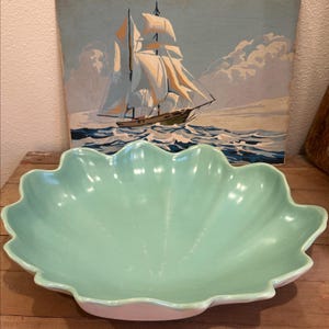 May include: A large, light green ceramic serving bowl with a scalloped edge. The bowl is set against a backdrop of a vintage painting of a sailboat on the ocean. The bowl's base is a soft pink.