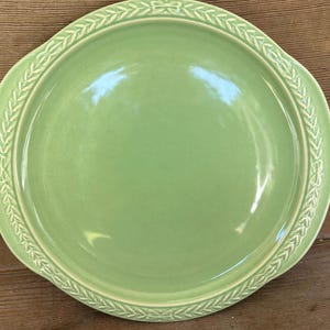 May include: A round, light green ceramic plate featuring a smooth, glossy center and a raised, textured border decorated with a repeating leaf and bow pattern in a lighter cream color. The plate has a wide rim and a simple, circular design.