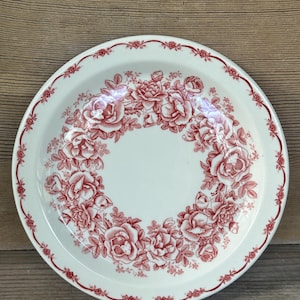 May include: A white ceramic plate with a red floral rose pattern. The plate features a ring of roses in the center and a decorative border. The plate is a classic design for serving food.