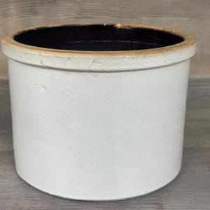 Peut inclure: Un pot en grès cylindrique blanc cassé avec un intérieur brun foncé et un bord brun clair. Le récipient antique a un design simple et classique, adapté à diverses utilisations. L'extérieur a une surface légèrement texturée.