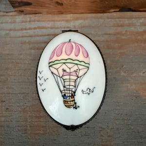 May include: An oval-shaped white trinket box with a hinged metal rim. The lid features a hand-painted illustration of a hot air balloon with a pink canopy, a brown basket, and two small figures inside, accompanied by birds in flight.
