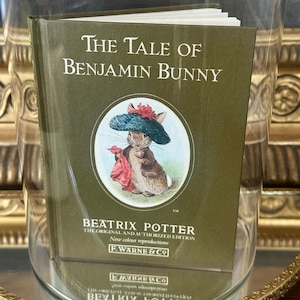 May include: A small hardcover book titled The Tale of Benjamin Bunny by Beatrix Potter. The olive green cover features a circular illustration of a rabbit wearing a blue hat and holding a red cloth, with white text and the F. Warne & Co logo.