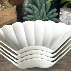May include: A stack of five white, scalloped-edge serving bowls. The bowls are arranged in a fan shape, with the top bowl in focus. The bowls are made of a smooth, glossy material. The background includes plants and a wooden crate.