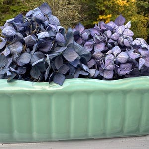 May include: A rectangular, pale green ceramic planter filled with dried blue and purple hydrangea flowers. The planter has a ribbed design and sits on a white surface, with a blurred outdoor background.