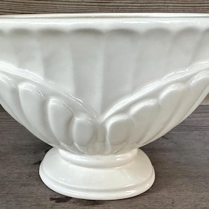 May include: A cream-coloured ceramic planter with a pedestal base. The planter features a wide, fluted rim and a decorative, leaf-like design around the body. The overall shape is an urn style, suitable for flowers or plants.