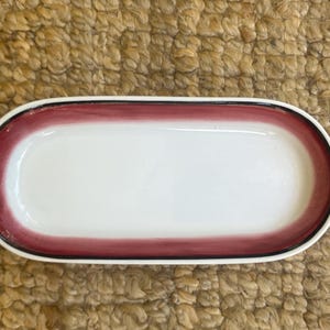 May include: An oval-shaped ceramic tray with a smooth white center and a wide, hand-painted deep red border. The outer edge of the rim is finished with a thin, distinct black line, creating a clean contrast against the white basin and red band.