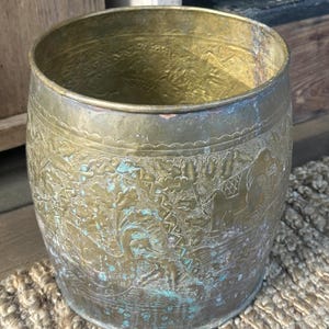 May include: A cylindrical brass container with etched designs. The container has a weathered appearance with areas of green oxidation. The etched patterns include figures and floral motifs. The container is sitting on a woven mat.