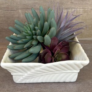 May include: A white square ceramic planter with a raised leaf pattern on the exterior sides. The planter contains a cluster of artificial succulents, including a large sage green plant, a smaller dark purple plant, and a spiky purple plant.