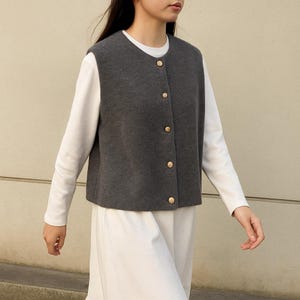 Può includere: Un gilet grigio antracite con bottoni dorati. Indossato sopra una camicia bianca a maniche lunghe e una gonna bianca. Il gilet ha uno scollo tondo e una vestibilità su misura. I bottoni sono uniformemente distanziati sul davanti.